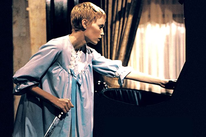 A woman in a nightgown holds a knife, leaning toward a crib in a dimly lit room with curtains.