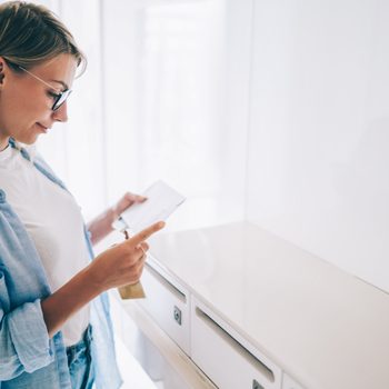 A person reads a letter while standing beside white mailboxes in a bright room.