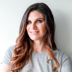 A person, smiling slightly, stands against a plain white background, wearing a light gray shirt with long brown hair cascading over their shoulders.