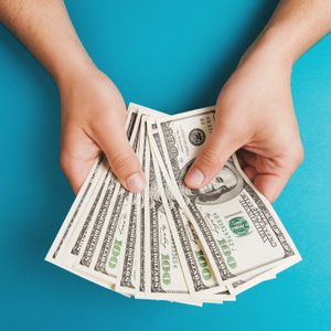 Hands holding several 100-dollar bills against a solid blue background.