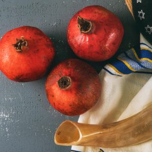 Three pomegranates rest on a gray surface beside a folded blue-striped cloth, shofar, and a black cap with embroidered stars.
