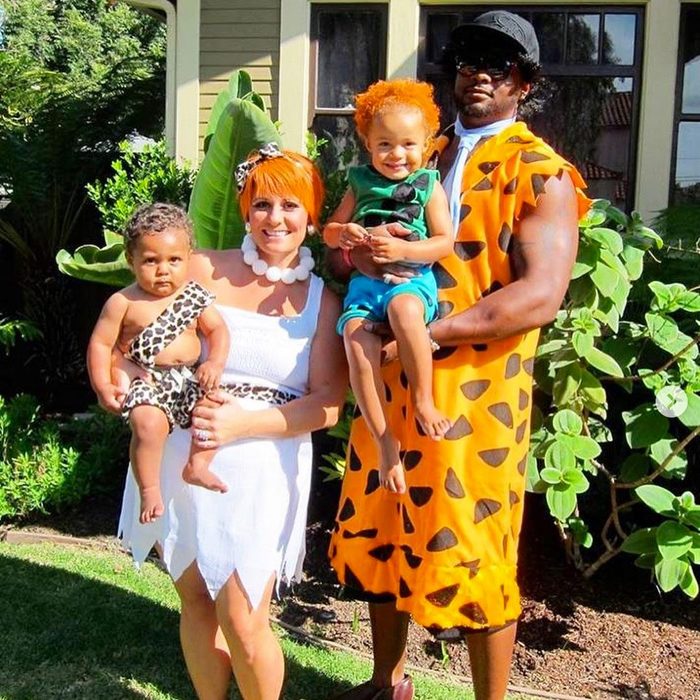 Family posing in Flintstones-themed costumes, standing outside a house with lush green plants and a sunlit lawn.