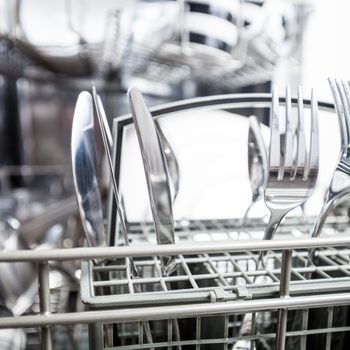 Silverware standing upright in a dishwasher basket, surrounded by racks, ready for washing.