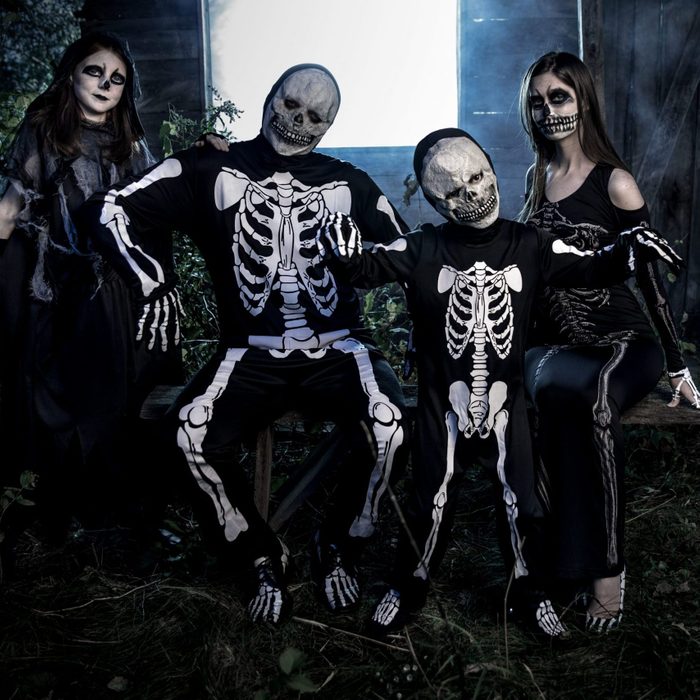People dressed as skeletons pose together outdoors on a wooden bench, surrounded by dark, leafy vegetation and illuminated by dim lighting.