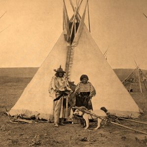 Teepee stands in open field; two individuals in traditional attire pose with a saddled dog, surrounded by more teepees.