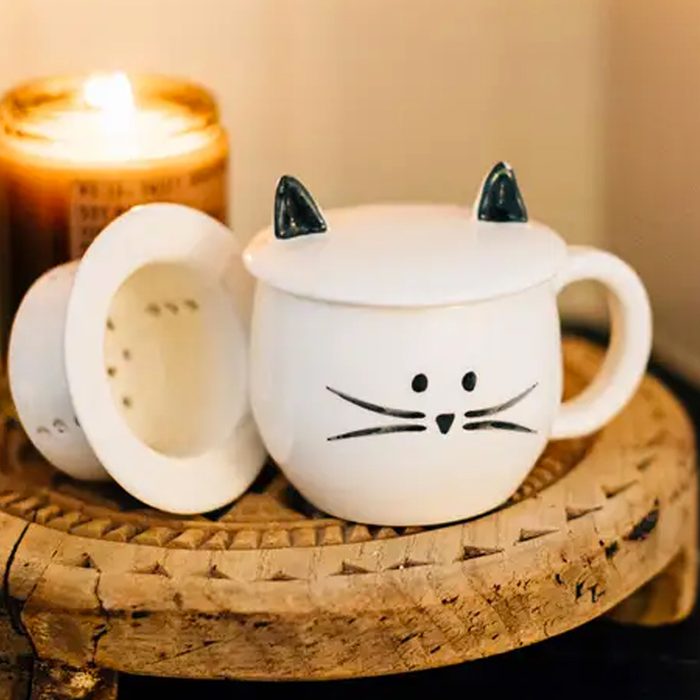 A cat-themed mug with lid rests on a carved wooden surface; a lit candle glows beside it.
