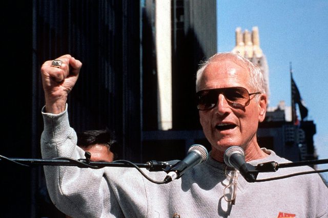 Man gestures passionately, speaking into microphones amidst a crowd, wearing sunglasses and a sweatshirt in an urban setting with tall buildings around.