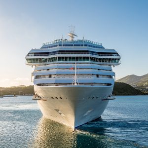 Cruise ship sails forward on tranquil water, surrounded by lush green hills under a clear blue sky.