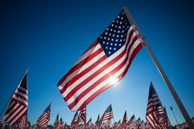 A prominent American flag waves under a clear blue sky, surrounded by numerous smaller flags in the background.