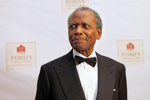 Man in tuxedo smiling slightly, standing against a backdrop with "Ford's Theatre" logos in a formal event setting.