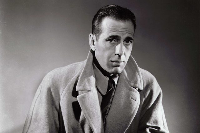 Man wearing a trench coat, gazing intently, sits against a plain, shaded background.