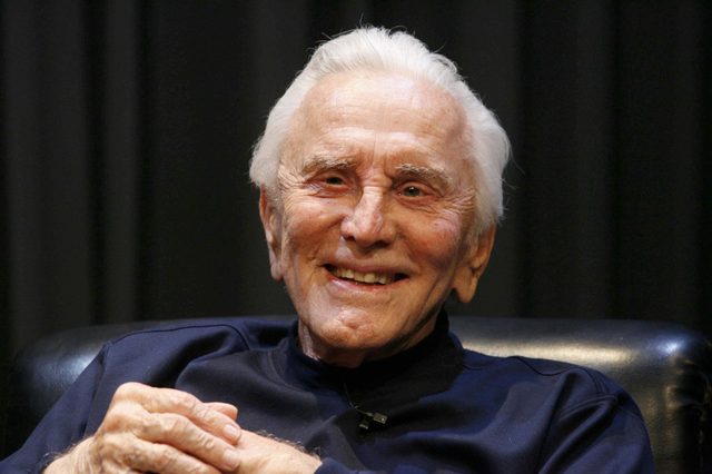 Elderly man smiling, seated in a leather chair, wearing a dark shirt against a dark curtain backdrop.