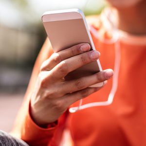 A person holds a smartphone, wearing headphones in an outdoor setting, dressed in an orange shirt.