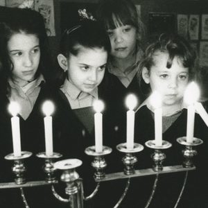 Children observe candles on a menorah, one child holding a candle, in a classroom decorated with artwork.