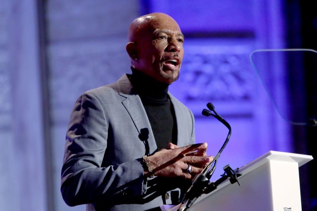 A person speaks at a podium with microphones, wearing a suit and turtleneck, against a blue-lit backdrop.