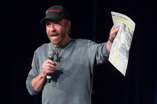Person holds a microphone and newspaper, gesturing energetically in a dark setting, wearing a cap and casual clothes.