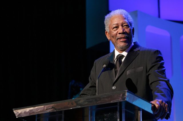 A man in a suit smiles while speaking at a podium, standing against a backdrop of blue and black geometric shapes.