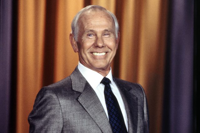 Smiling man stands in a suit with a patterned tie, in front of gold and blue curtains.