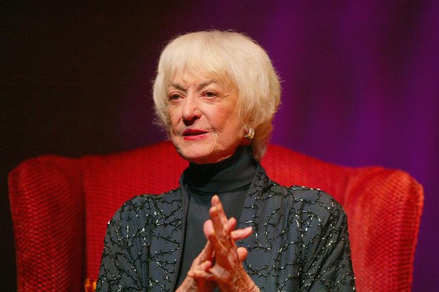 An elderly woman sits with clasped hands in a dark, ornate jacket against a red armchair, set in a dark, softly lit environment.