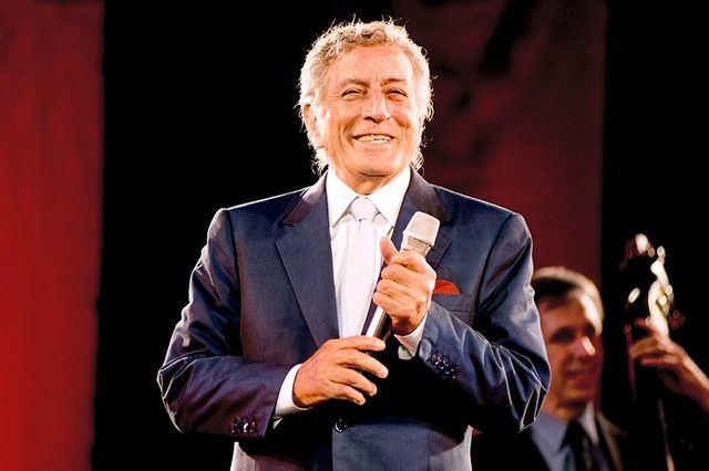 Man in a suit holds a microphone, smiling on stage with a musician in the background against a dark backdrop.