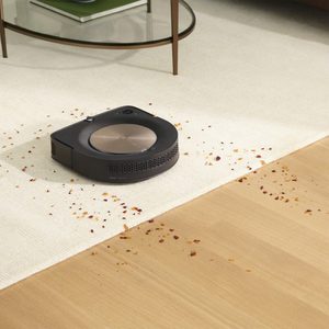 Robot vacuum cleaning crumbs between a carpet and wooden floor; a glass table with books is in the background.