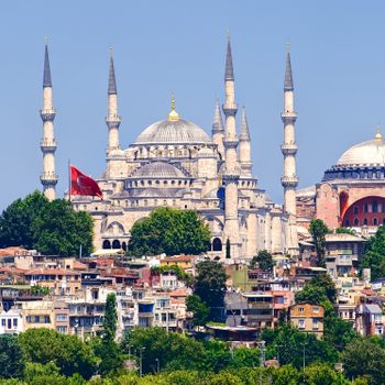 Domed mosque towers above colorful city buildings, surrounded by minarets and greenery under a clear blue sky. A red flag waves nearby.