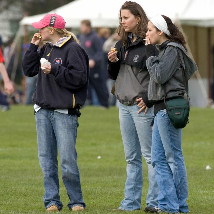 Three people stand eating on grass near tents, wearing casual clothes and jeans, with others in the background.