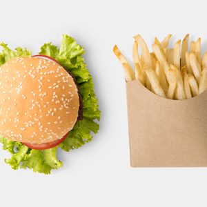 Burger with lettuce and tomato sits beside carton of fries on a white background.