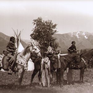 Three individuals ride decorated horses, standing still in a field, with a teepee and snow-capped mountains visible in the background.