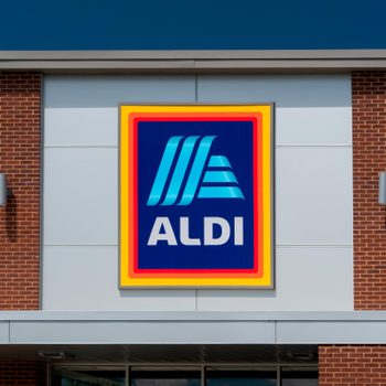 Sign displaying "ALDI" is mounted on a building facade, featuring brick and metal elements, against a clear blue sky backdrop.