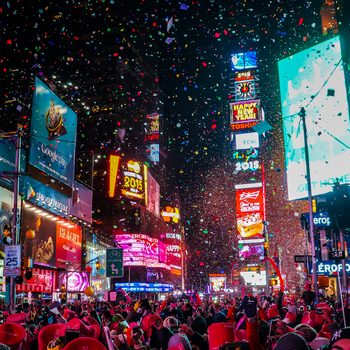 Confetti falls over a crowded celebration in Times Square; vibrant billboards illuminate the scene, including a New Yearâ€™s Eve countdown.