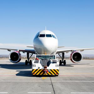 Airplane being towed by a pushback vehicle on a sunny airport tarmac, with clear blue sky and distant runway in the background.