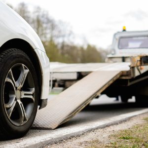 Car approaches ramp, ascending a flatbed tow truck on the roadside, surrounded by trees and grass.
