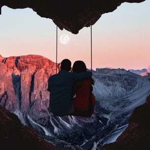 A couple sits on a swing framed by a heart-shaped cave, overlooking a stunning sunset mountain landscape with a full moon.