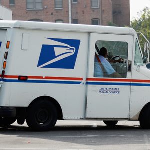 A USPS mail truck drives through an urban parking area, featuring a postal logo and blue, white, red stripes.