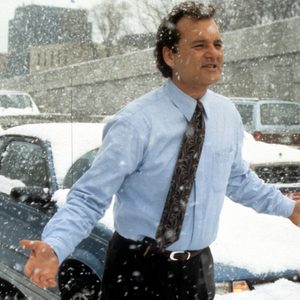 Man stands with arms outstretched, expressing confusion, in snowy traffic jam, surrounded by cars and falling snowflakes.
