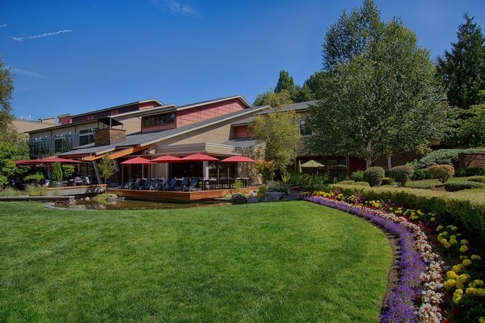 House features red umbrellas over patio chairs, surrounded by lush greenery and colorful flowerbeds in a sunny, landscaped garden.