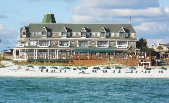 Large beachfront building stands, featuring green roofs, overlooking sandy beach with lined umbrellas and ocean waves under a partly cloudy sky.