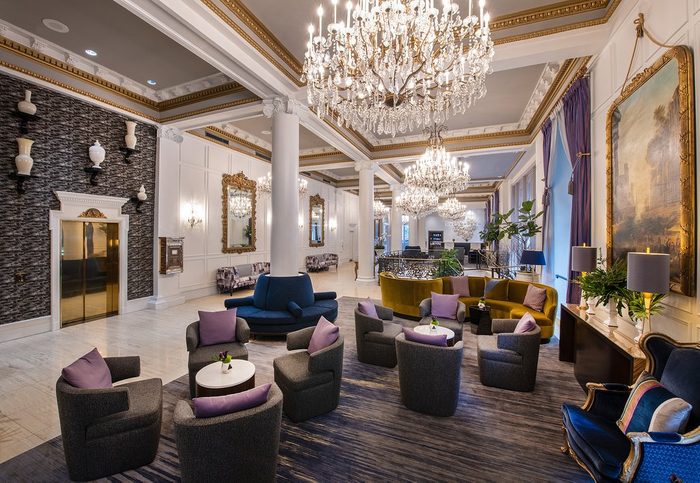 Elegant lobby features plush seating arranged around tables, illuminated by chandeliers, with ornate mirrors, large artworks, and pillars enhancing the classic decor.