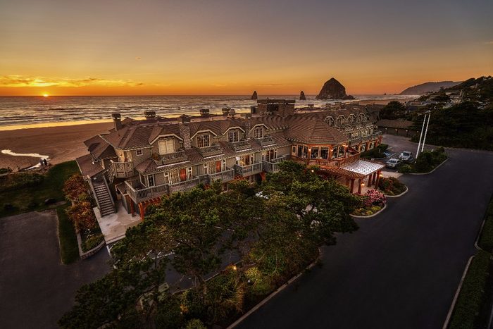 Building overlooks ocean during sunset, surrounded by gardens, with visible haystack rocks in the distance.
