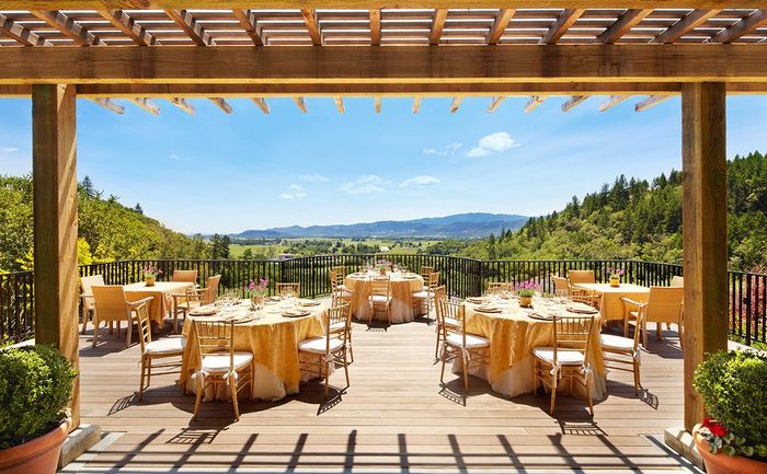 Tables with golden cloths stand ready on a wooden patio under a pergola, overlooking expansive green hills and a clear blue sky.
