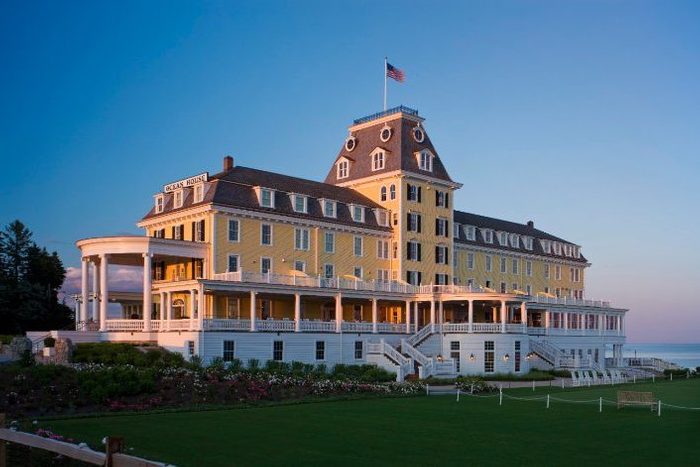 A large, yellow Victorian-style hotel stands illuminated at sunset, with a landscaped garden and view of the ocean. The sign reads 