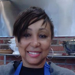 A person smiles while wearing hoop earrings and an apron. They are in a kitchen setting with brick walls and metal cookware.