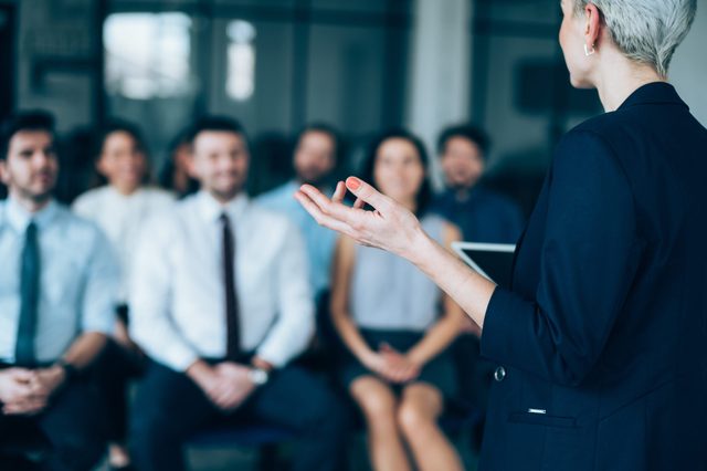 A speaker gestures while giving a presentation, addressing an attentive group of people in a formal setting.