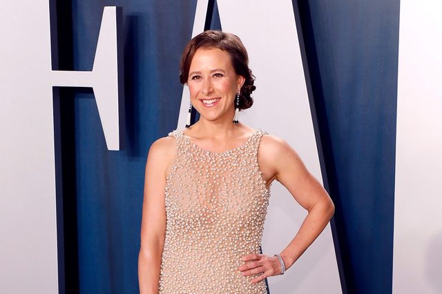 A person stands smiling, wearing a pearl-covered dress, in front of a blue and white backdrop at an event.