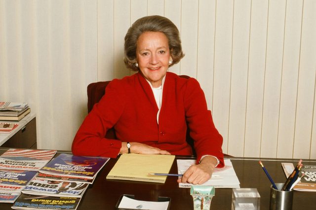A smiling woman in a red jacket sits at a desk with magazines, papers, and a pencil holder, in an office with vertical blinds.
