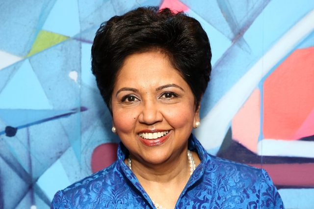 A person smiling, wearing a blue patterned jacket and pearl necklace, stands in front of a colorful geometric background.