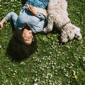 Person lies on grass, smiling, beside a fluffy dog. Surrounded by small white flowers in a sunny outdoor setting.
