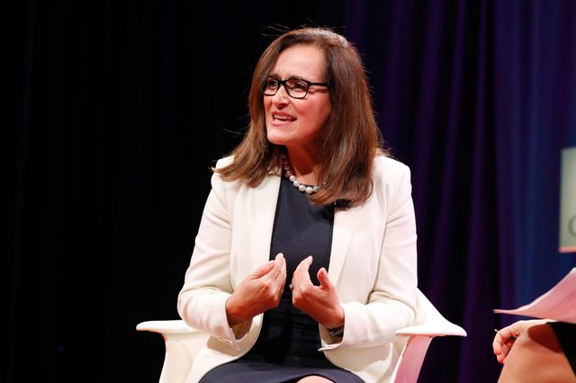A person gestures while speaking, sitting in a white chair on stage, wearing glasses and a white blazer.
