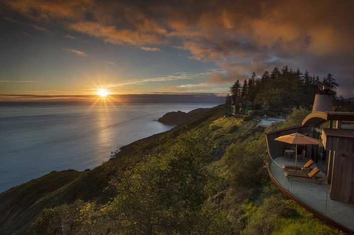 The sun sets over the ocean, illuminating a hillside with a house featuring a patio and umbrella, surrounded by trees and clouds.